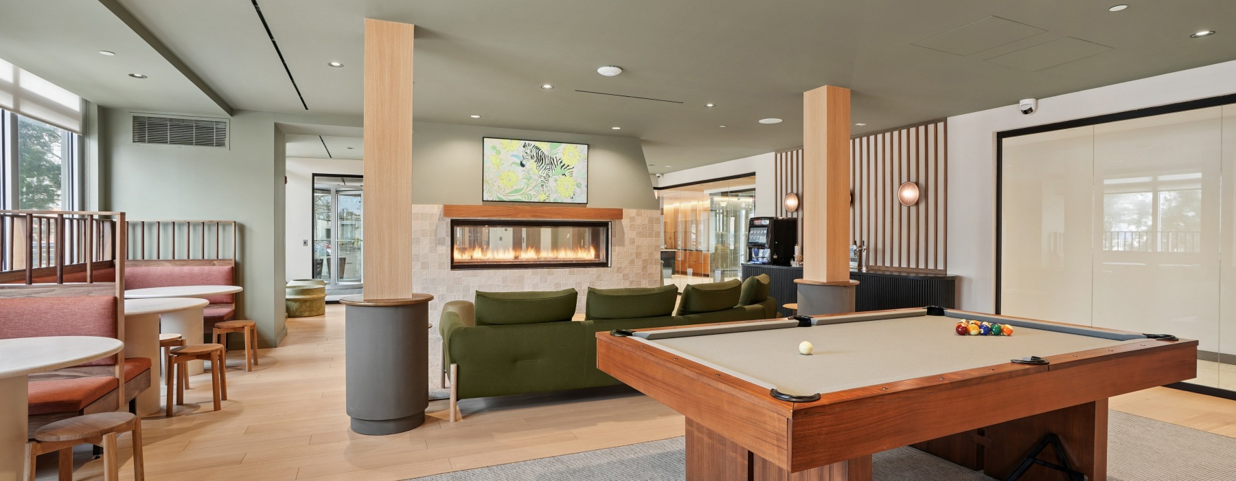 Clubroom at Atmark Apartments in Cambridge with a billards table, green couch in front of a fireplace, and row of tables with banquette seating in front of windows